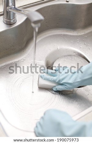 Gloved hands cleaning a stainless steel sink with a sponge.
