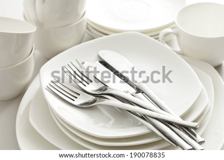 Silver forks and knives rest on a stack of plates and dishes, with white mugs behind.