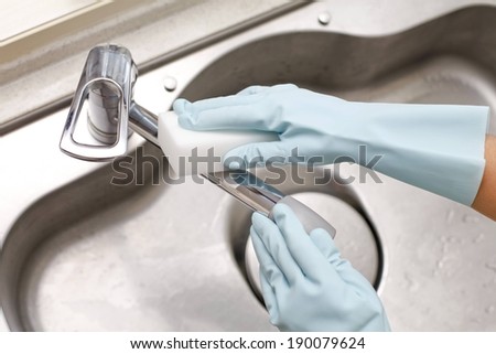 Hands in blue gloves scrub a steel sink faucet.