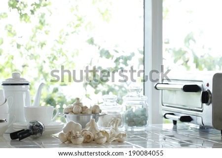 A white tile kitchen surface with a coffee pot, toaster oven, garlic and bowl of mushrooms.