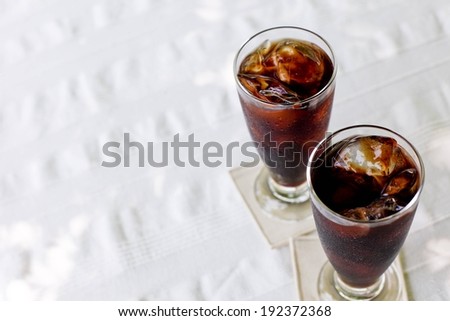 Two tall glasses filled with brown liquid and ice cubes on white coasters and tablecloth.