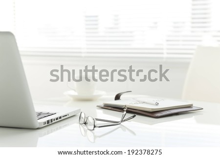 A desk, with a laptop, clipboard, coffee mug, and pair of glasses.