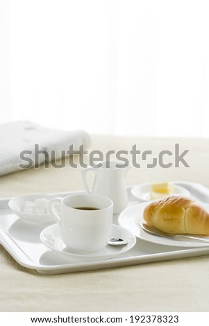 A white tray with coffee, croissant, cream pitcher and sugar cubes beside a newspaper.