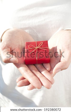 Two open hands holding a small gift box wrapped in red with a gold ribbon.