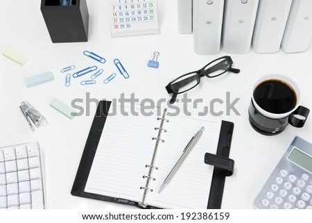 Coffee served in a black mug, glasses, planner and paperclips scattered on a desk.