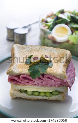 A sandwich with three slices of bread, meat, green stalks and cheese with green olive garnish.