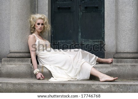 Pale model with big hair sitting against a cement column in a white dress