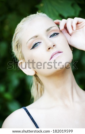 A gorgeous blond female model with wet hair and her hand on her face posing in a forest on a sunny day looking at the camera with green eyes.