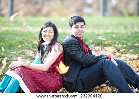 A young and happy Indian couple sitting on leaves back to back in the Fall on a cloudy day.