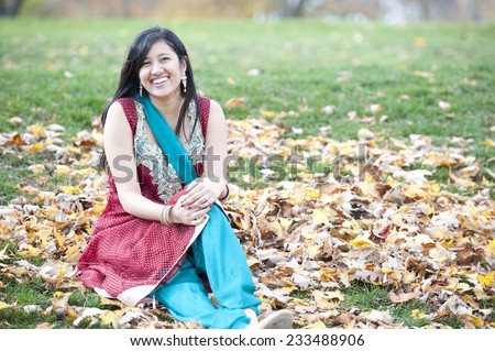 A young happy Indian girl sitting in a pile of leaves outdoors on a cloudy day.