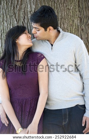 A young and happy Indian couple kissing with a tree background on a cloudy day.