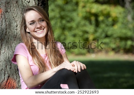 An attractive and happy young brunette girl sitting next to a tree on a sunny day.