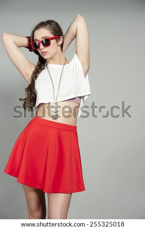 Attractive young brunette girl wearing a pair of red sunglasses, white tank top and a red mini skirt on a gray studio background.