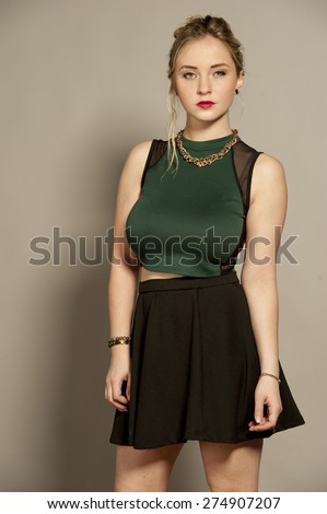 Beautiful busty young female brunette with straightened hair in a studio setting while wearing a green top and a short black mini skirt on a gray background.