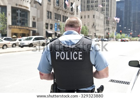 A Chicago city policeman standing on the road on a sunny day wearing a bullet proof vest mentioning Police on his body. On the background, cars and tall buildings are seen.