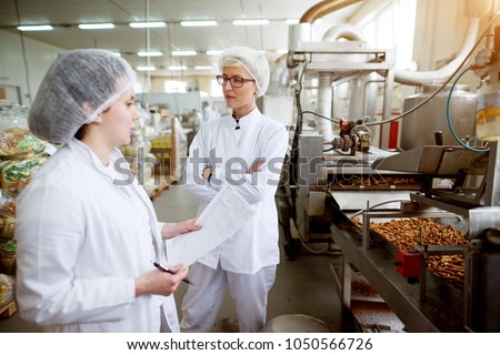 Focused dedicated tired female employee giving rapport in food production factory to her worried and annoyed superior while holding statistic notes and wearing sterile cloths.