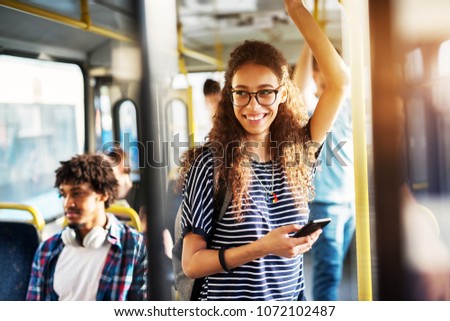 Young gorgeous cheerful woman is standing on the bus using the phone and smiling.