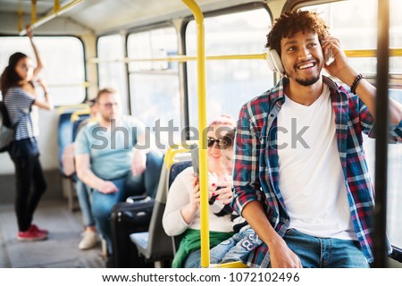 A group of people traveling by the bus with a young cheerful handsome man in the first plan of the picture who is listening to music on his headphones and smiling.