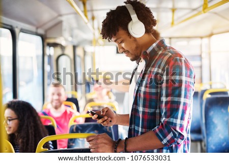 Young handsome man with a headset on is listening to music while looking at his phone and standing on the bus.