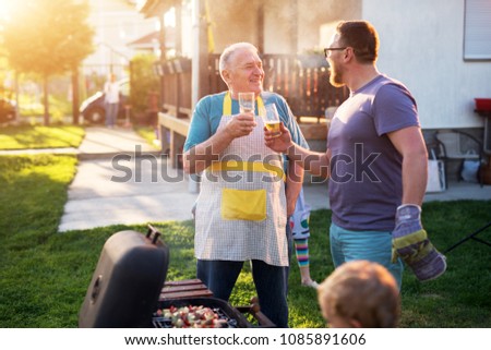 Elderly Father and mature son are saluting with the beer in front of the grill in their house backyard on a beautiful day.