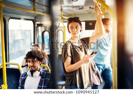 Young beautiful woman is standing in the bus using phone and holding onto the bar while waiting to arrive at her destination.