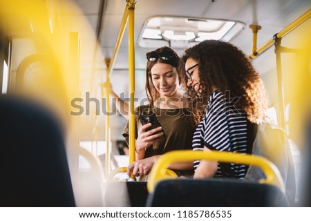 Two best young sweet girl friends standing in a bus and looking in a telephone.