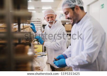 Picture of two male food factory employees in sterile clothes packing fresh made cookies and having fun.