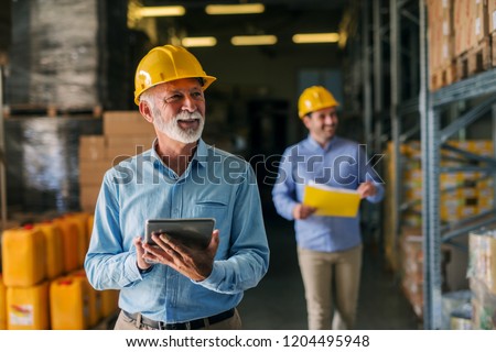 Picture of father and son walking through their warehouse with helmets on their heads. Father is holding digital tablet in his hand and looking at shelf full of boxes with smile on his face.