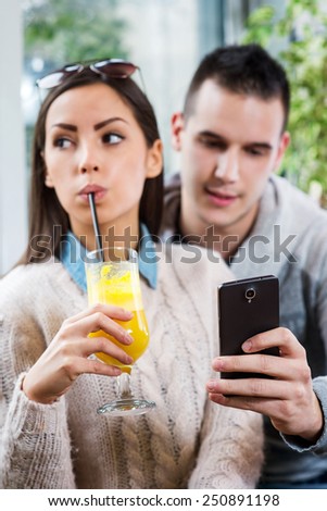 Young beautiful couple sitting in cafe. Boyfriend showing her a smart phone, she is not interested.