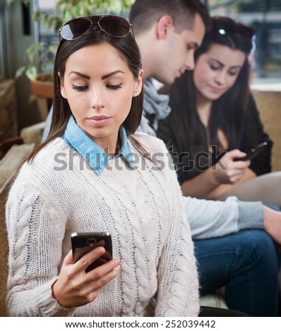 Young female sitting in cafe and looking at smart phone, two people are blurred in background.
