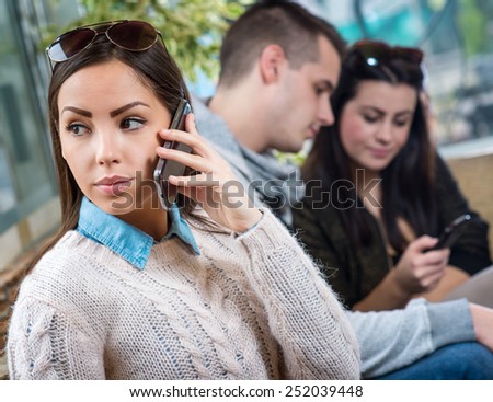 Young female sitting in cafe and talking on smart phone, two people are blurred in background.