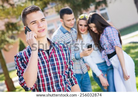 Portrait of young beautiful boy talking at smart phone, friends in background looking at smart phone. Enjoying the park in spring day. Teenagers.