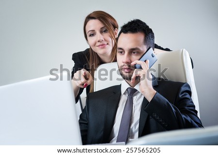 Handsome businessman sitting at office looking at lap top screen, talking at smart phone, while attractive business woman showing him something on lap top. Office, business and marketing concept.
