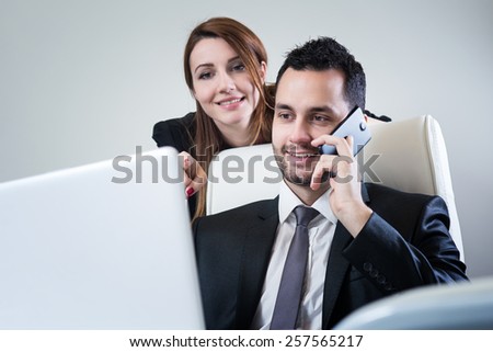 Handsome businessman sitting at office looking at lap top screen, talking at smart phone, while attractive business woman showing him something on lap top. Office, business and marketing concept.