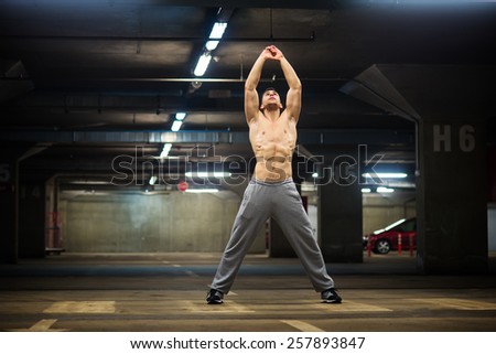 Handsome muscular young man stretching at parking garage, natural lights, dark place.