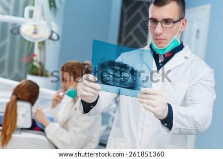 Dentist looking at roentgen, other female dentist examining a patient in background. Bright dentist office. Empty space for text.