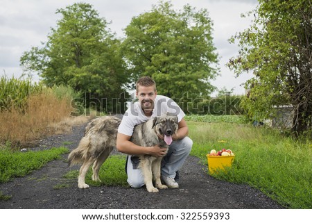 Man\'s best friend. Young man hugging a dog, both looking at camera. Bucket with fruit stands next to them.