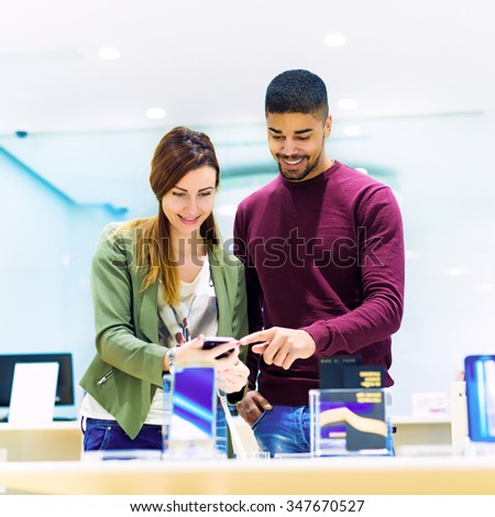 Woman is holding a mobile phone and black man is pointing finger at phone. They are looking for new smart phone. Mobile shop. Shallow depth of field.