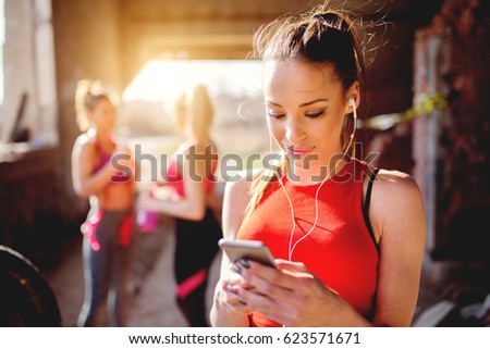 Young attractive fitness model listening music on smart phone charching positive energy before workout outside. Sun is shining and friends are behind preparing for training.