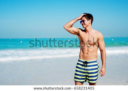 Handsome shirtless muscular fitness man at the beach, looking aside and smiling.