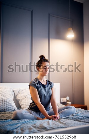 Peaceful pretty middle aged active woman practising yoga on the bed holding eyes closed, sitting in Half Lotus exercise, Ardha Padmasana pose and wearing sportswear at night.