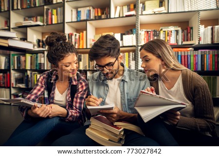 Concept of education, library, students and teamwork. Three charming focused productive young studying friends sitting on the floor in the library and learning together from notes and the books.