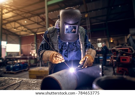 Close up portrait view of professional mask protected welder man in uniform working on the metal sculpture at the table in the industrial fabric workshop in front of few other workers.