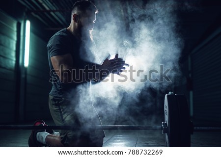 Focussed hardworking active fitness strong muscular bearded bodybuilder man clapping hands with chalk powder before strength training with a heavyweight dumbbell in the underground garage.