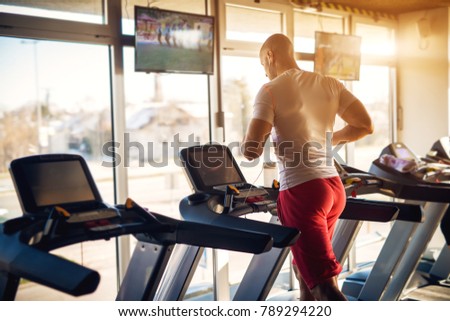 Rear view of strong motivated and focused muscular bald bodybuilder man running on the treadmill with earphones in the modern sunny gym with tv in front.