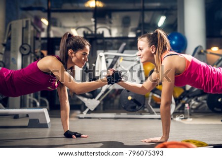 Two young motivated aggressive attractive focused sporty active girls doing push ups and holding hands together while looking each other in the modern gym.