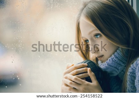 Portrait of a sad Teenager Girl with long blonde Hair. She is sitting on a Window in the Rainy  Day