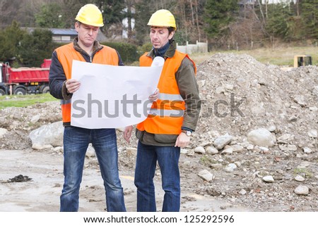 Project manager and architect are standing in a construction site looking in a construction plan Project manager and architect are standing in a construction site looking in a construction plan