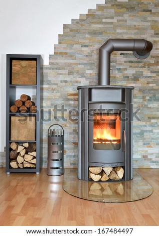 Wood Fired Stove Burning With Fire-Wood. Rack With Fire-Wood, Fire-Irons And Briquettes In Front Of Brick Wall