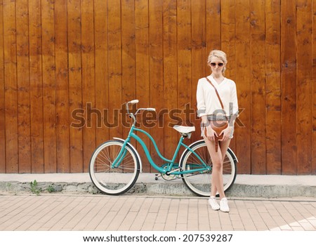Young sexy blonde girl is standing near the vintage green bicycle with brown vintage bag in orange sunglasses.